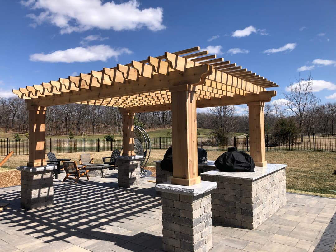 Wooden pergola with stone pillars and barbecue grills in a landscaped backyard.