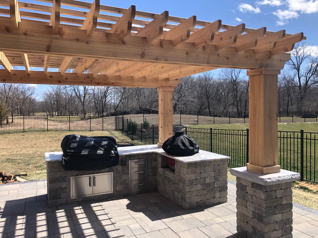 Outdoor kitchen with a pergola, stone countertops, and grills in a spacious backyard setting.