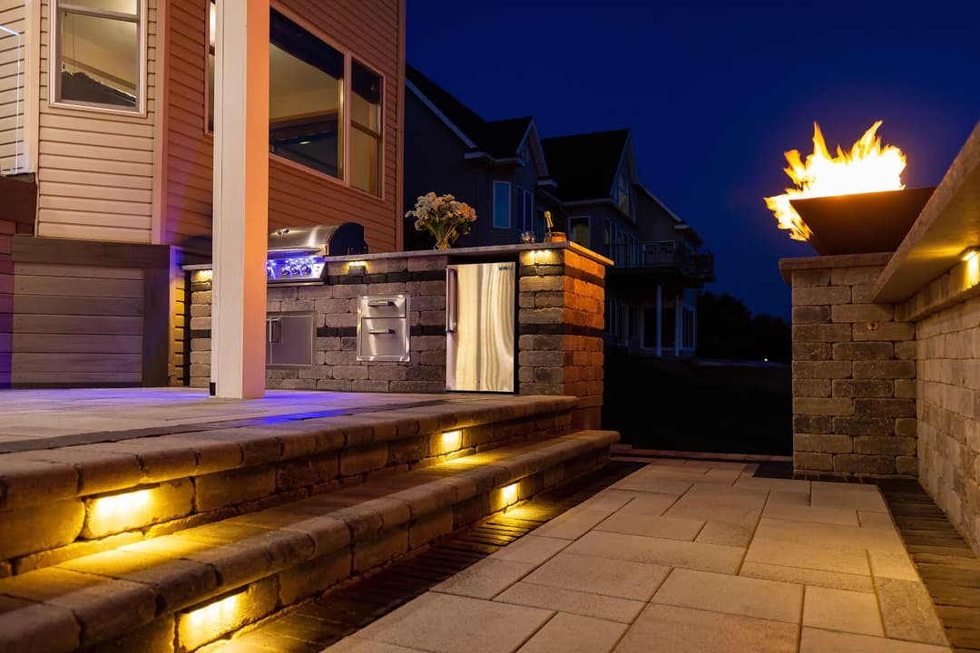 Modern outdoor kitchen with grill and fire feature, illuminated at night. Elegant stone pathway.