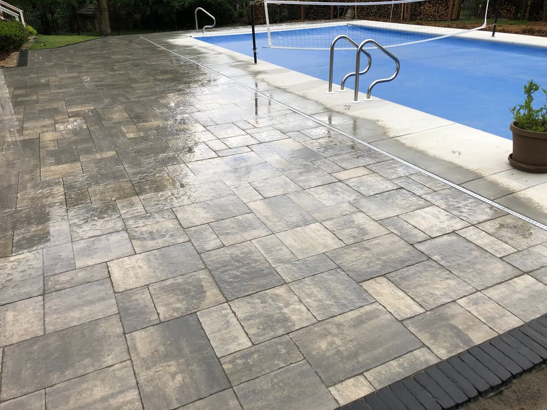 Wet paver patio beside a swimming pool with a net, surrounded by greenery.