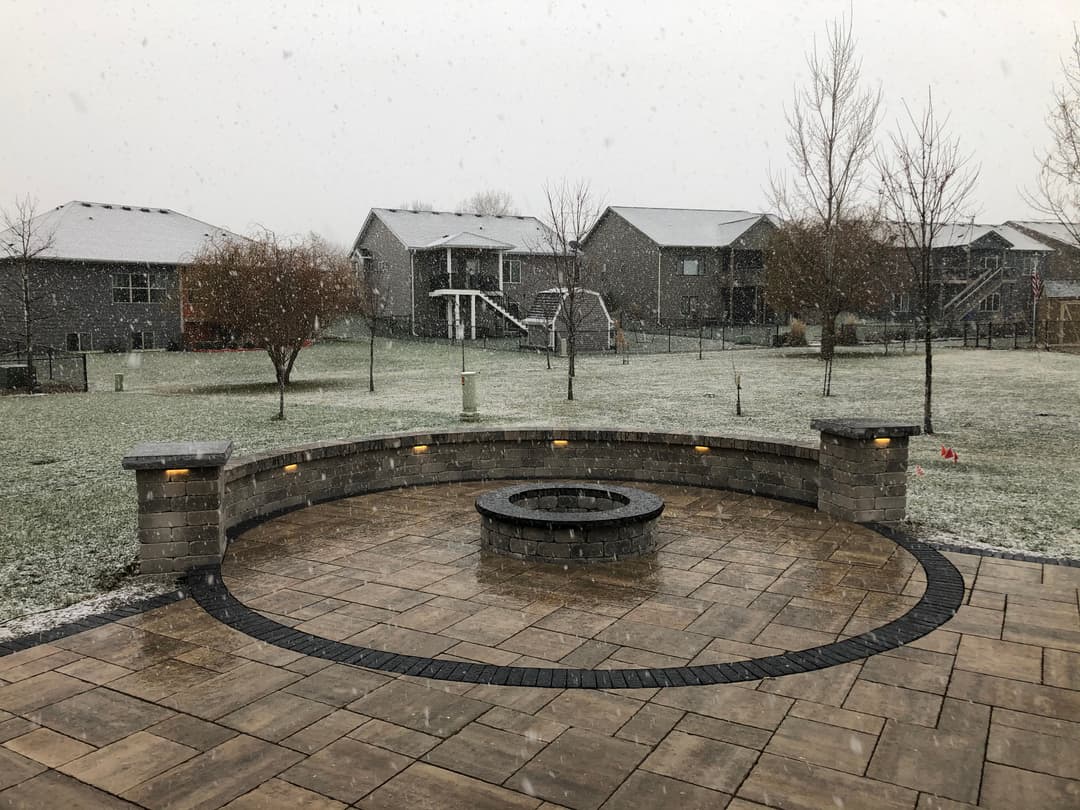 Winter backyard scene with snow falling over circular stone firepit and residential homes.