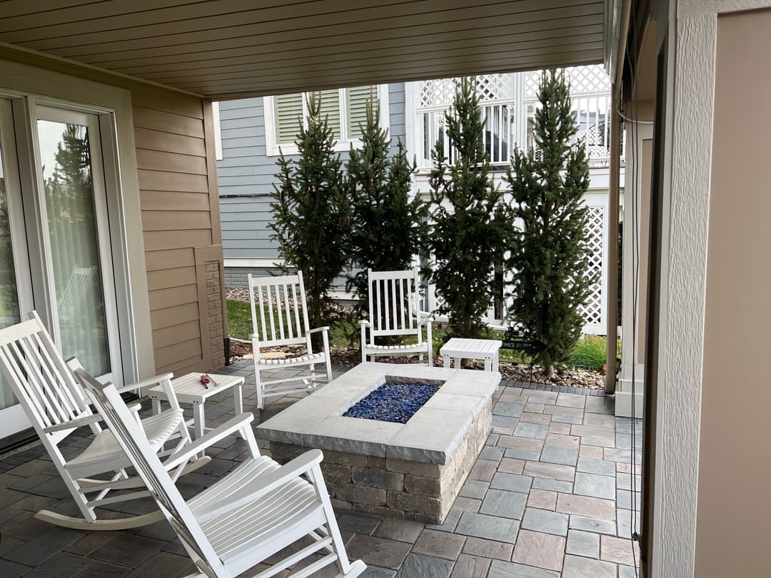 Outdoor seating area with white rocking chairs around a stone fire pit, surrounded by greenery.