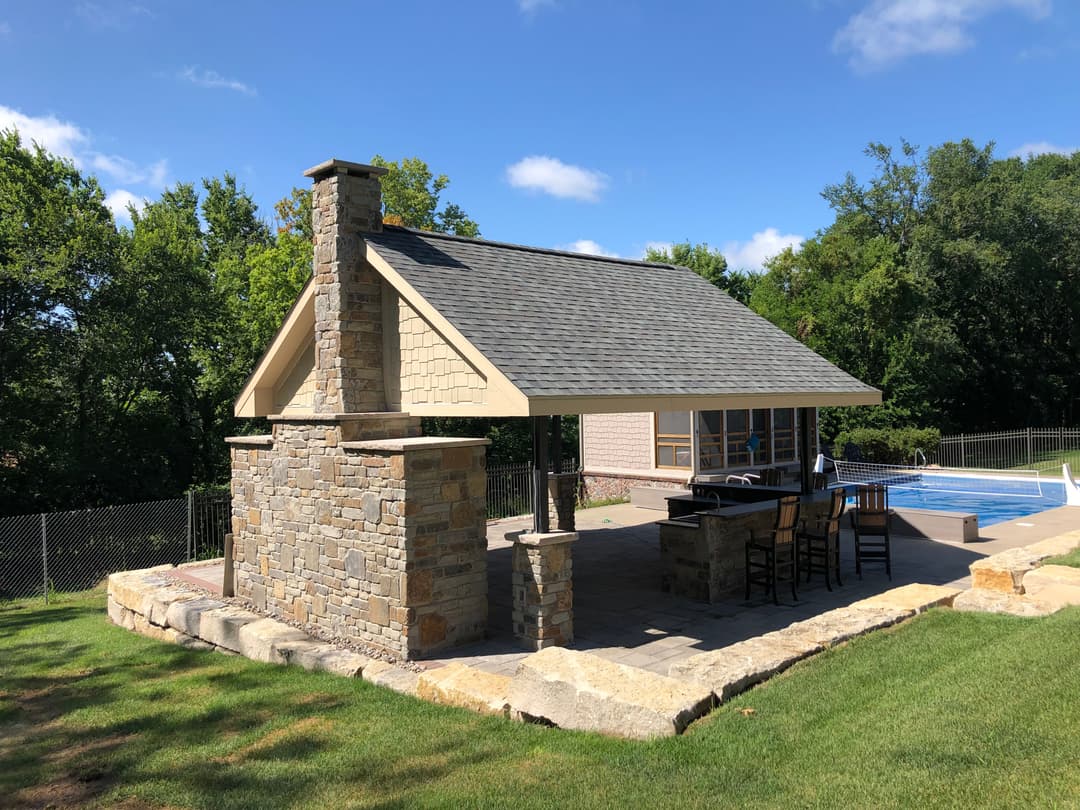 Outdoor poolside pavilion with stone fireplace and bar area surrounded by greenery.