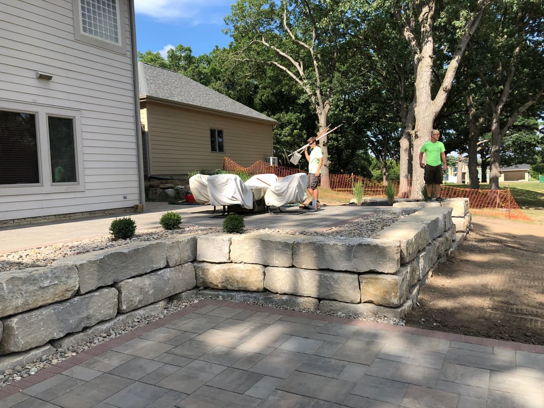 Workers landscaping a stone patio with a home in the background and trees overhead.