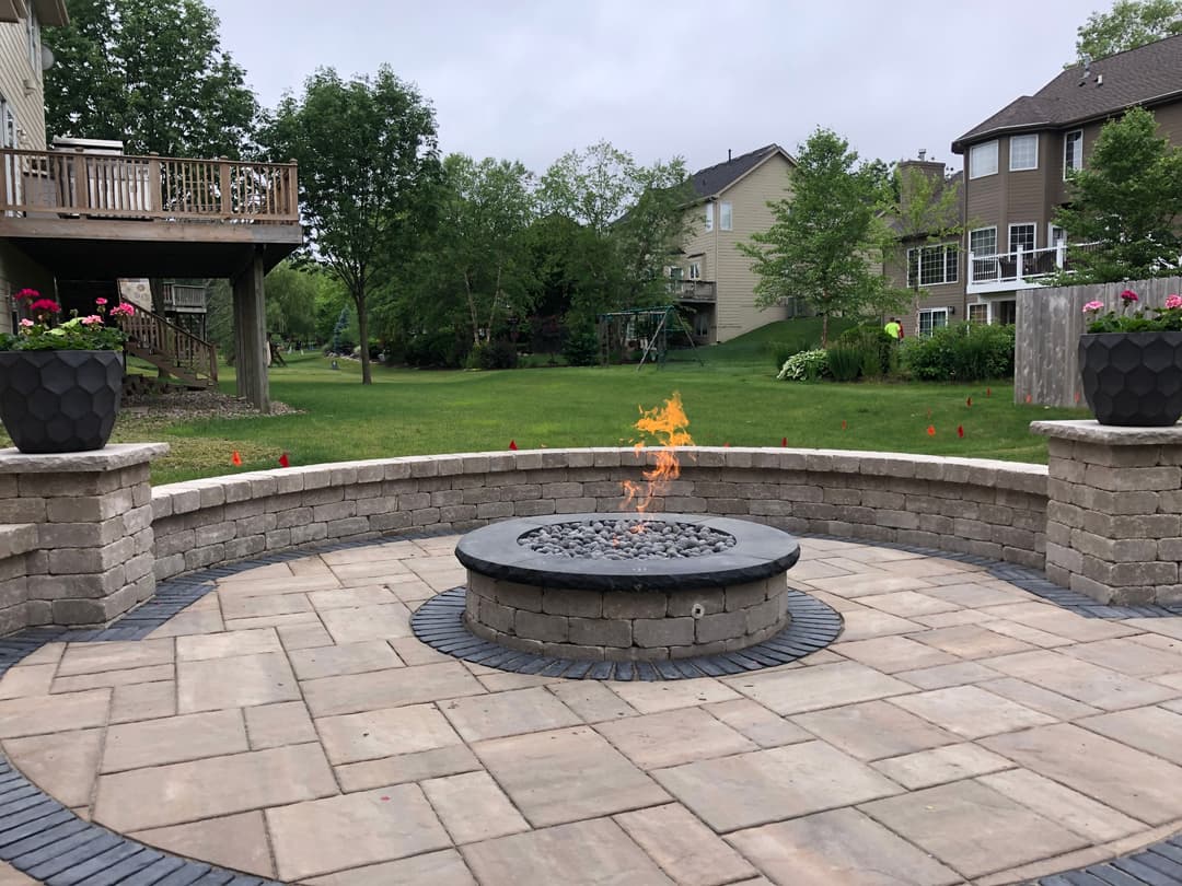 Modern outdoor fire pit with seating, surrounded by a lush green yard and landscaped flower pots.