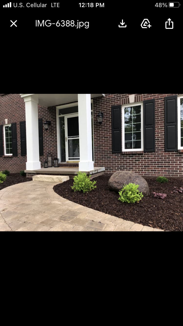 Front porch of a brick house with black shutters, landscaped garden, and decorative boulders.