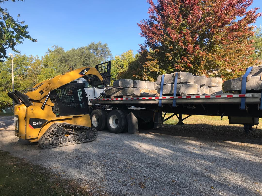Yellow skid steer loader unloading large stones from a flatbed truck in a residential area.