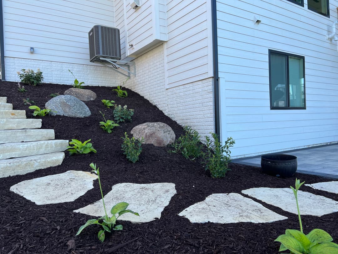 Landscape design featuring stone steps, boulders, and green plants alongside a modern home.