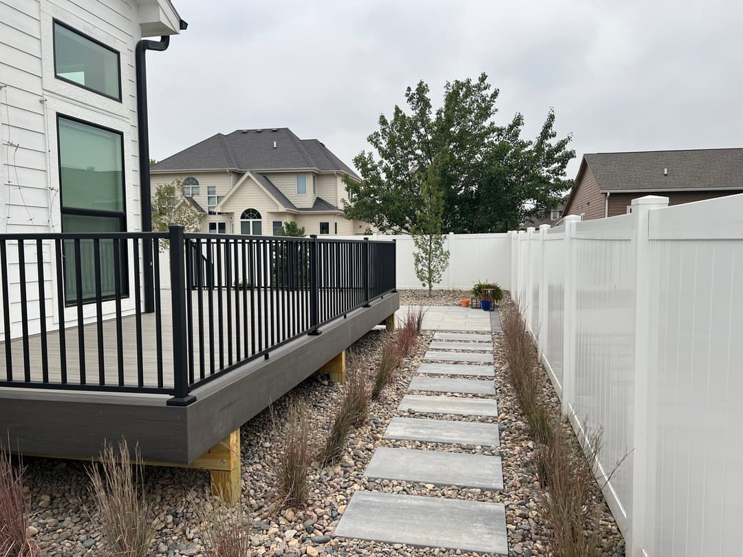 Modern backyard with a wooden deck, stone pathway, and landscaped garden surrounded by a white fence.