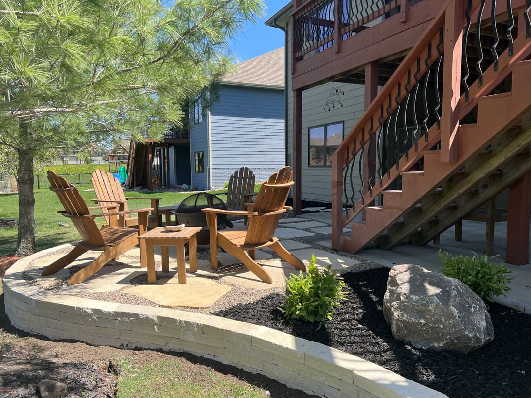 Cozy outdoor seating area with wooden chairs, stone patio, and landscaped garden under sunny sky.