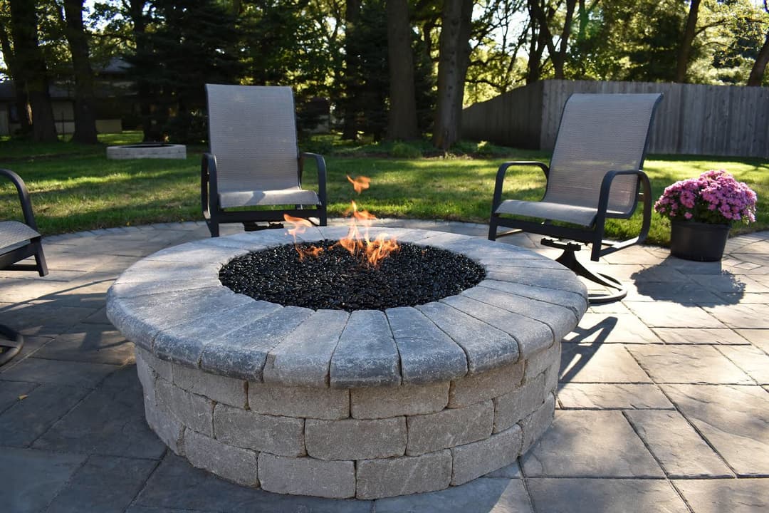 Outdoor fire pit with black glass stones, surrounded by two chairs and a blooming flower pot.