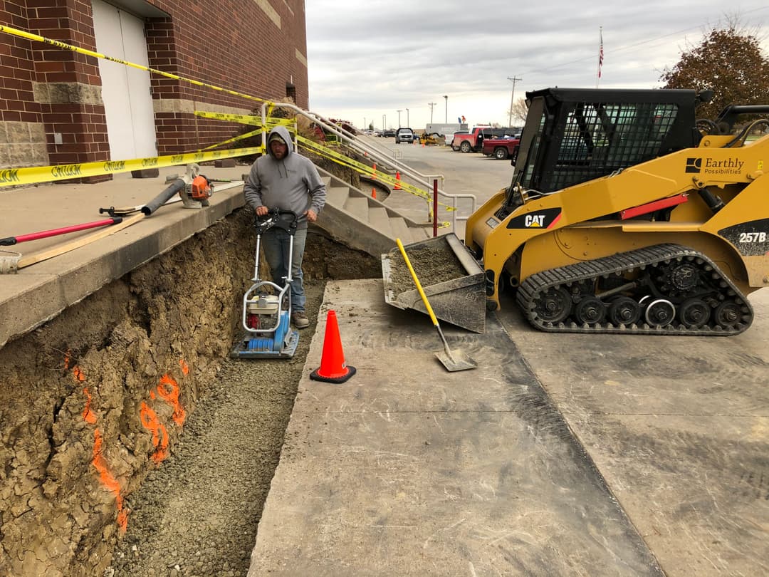 Construction worker using a plate compactor on a job site with heavy machinery and safety cones.