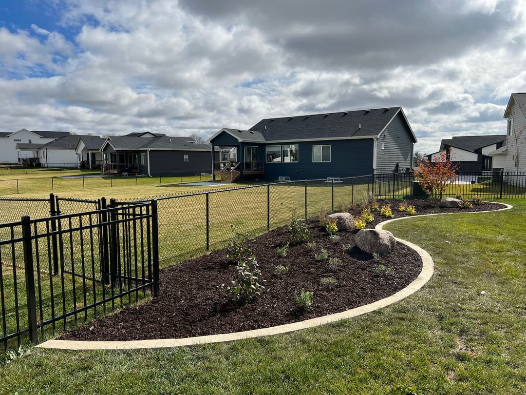 Residential backyard landscaping with curved garden bed, fence, and modern homes in the background.