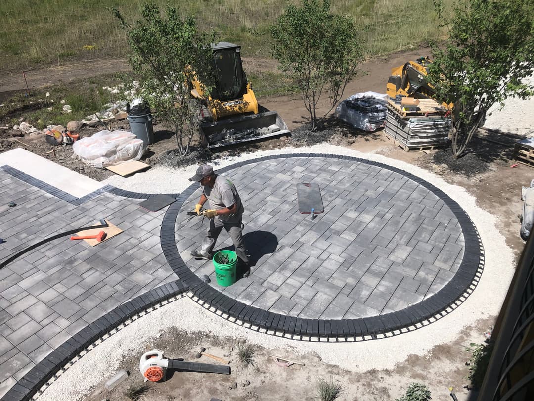 Worker installing gray pavers in a circular outdoor patio with landscaping materials nearby.