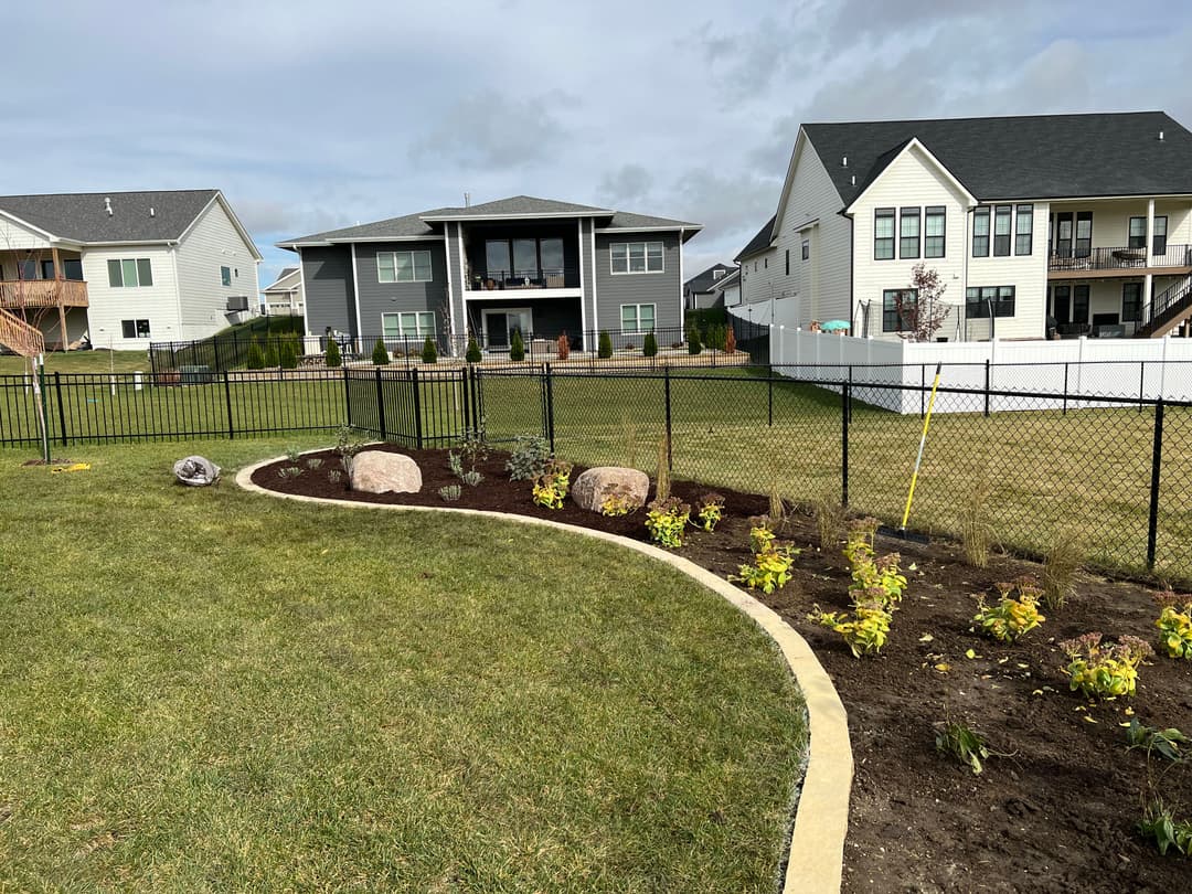 Lush backyard garden with landscaped flowers, rocks, and a view of houses and a fence.