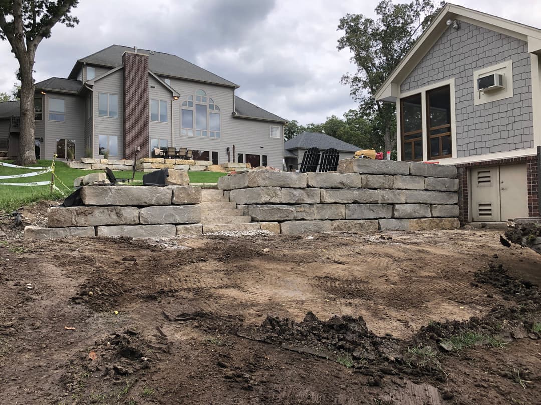 Stone retaining wall under construction beside a large modern home with a grey exterior.